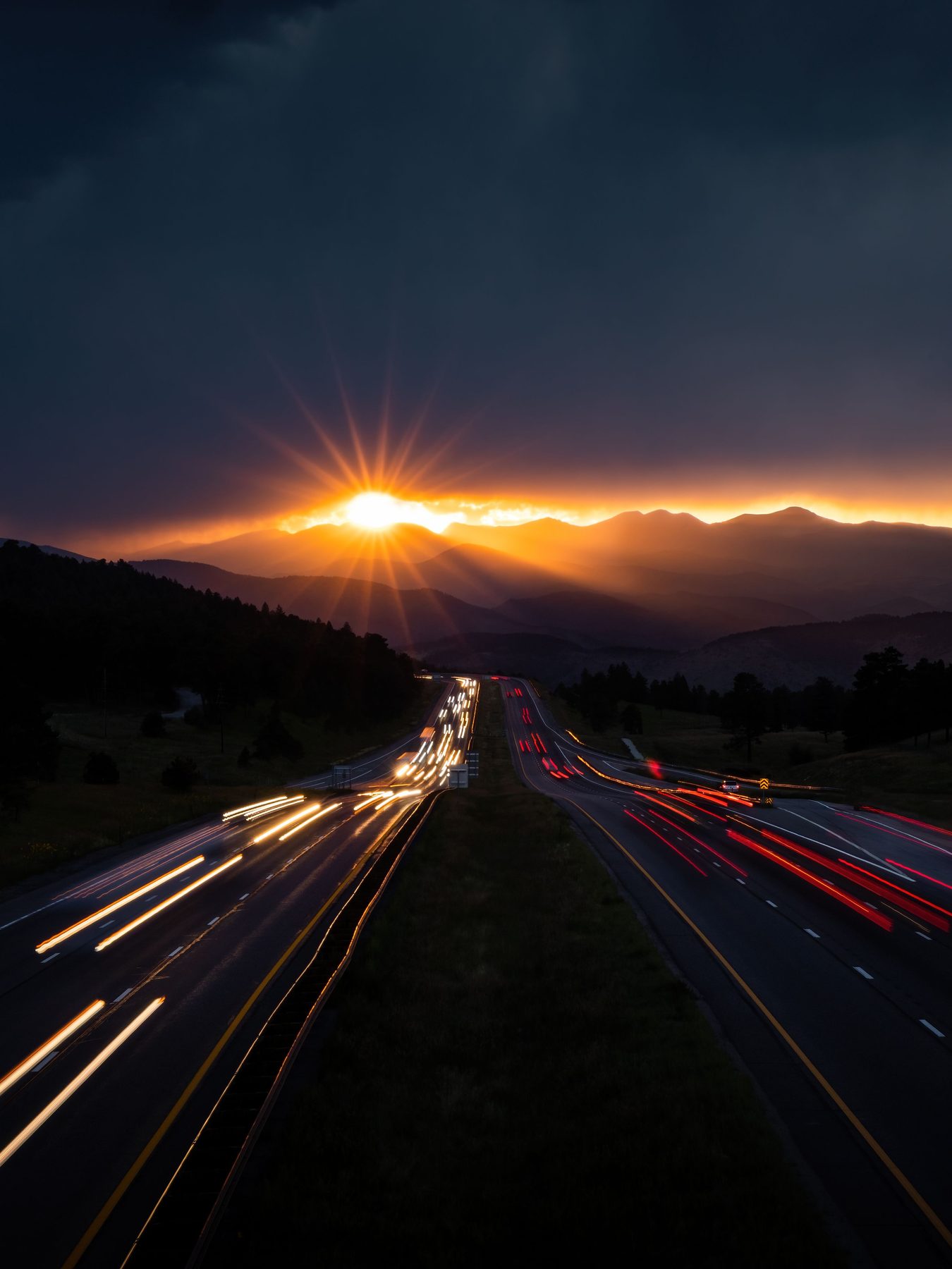 A divided highway at dusk, headlight and taillight trails running toward a sun setting behind distant ridges