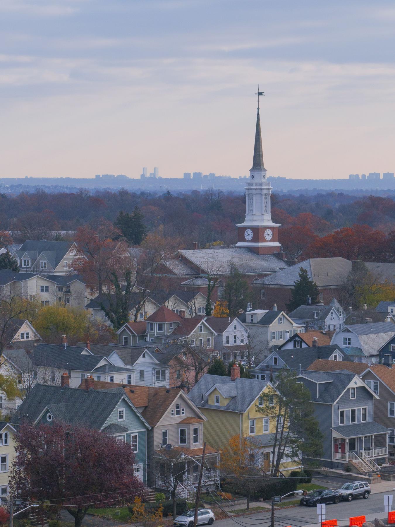 Rooftops of a New Jersey town at dusk, a clock-tower steeple rising above the houses with a distant skyline on the horizon